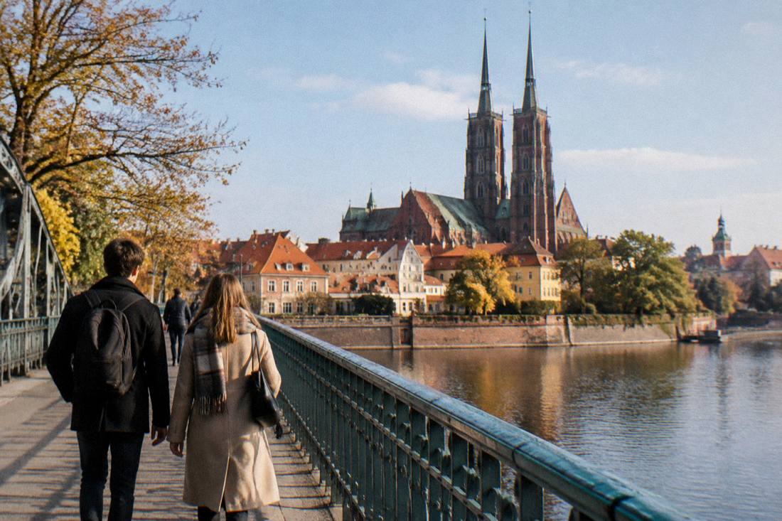Two people walking light-handed across a bridge over a river with a Gothic cathedral behind