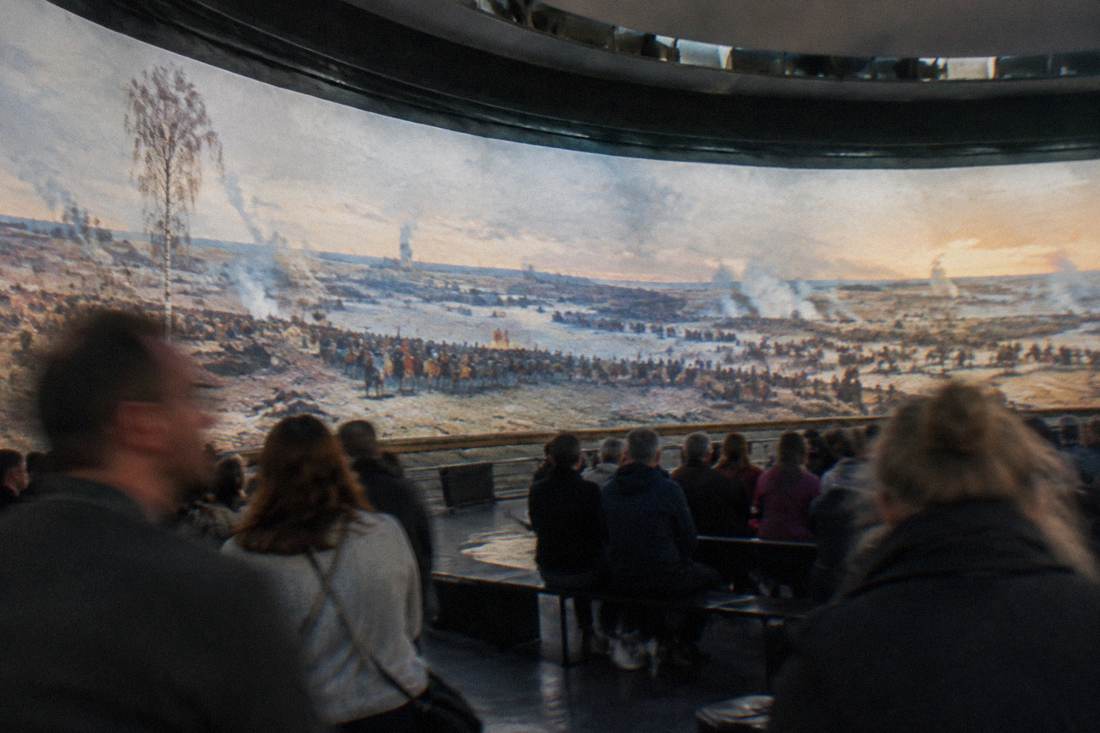 A seated crowd inside a circular gallery viewing a large panoramic painting in Wroclaw.