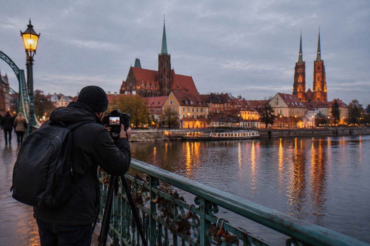 A photographer standing on a Wroclaw bridge at dusk with camera on a tripod, the illuminated Odra river and old town behind