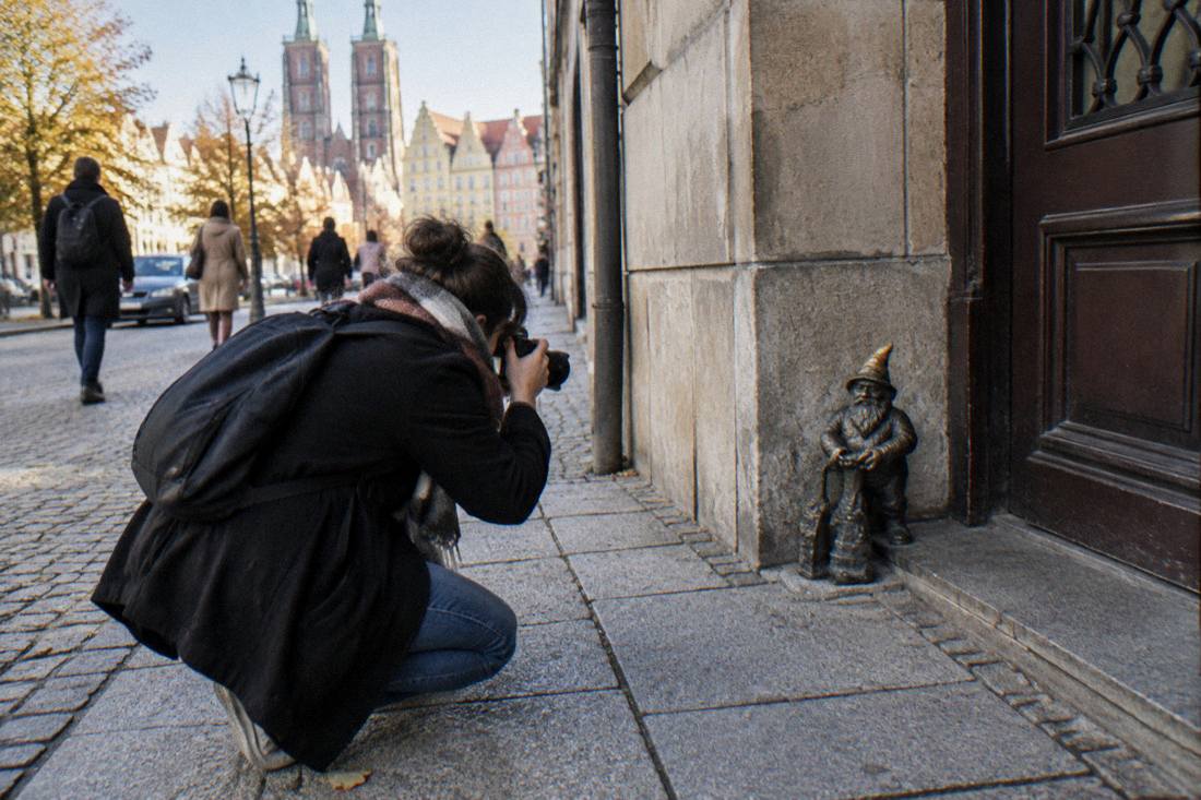 A photographer crouching on a Wroclaw pavement to capture a small bronze dwarf statue beside a doorway