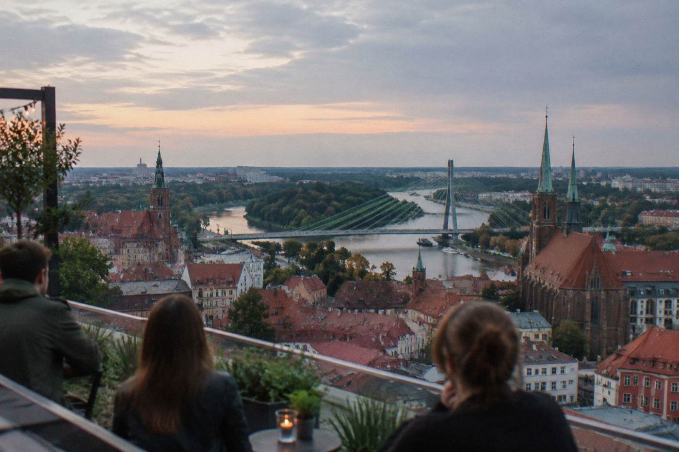 Evening view over red rooftops and church towers of Wroclaw from a rooftop terrace, with the Odra river visible in the distance.
