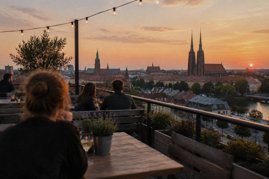 A rooftop terrace at dusk in Wroclaw with string lights, wooden tables and a view over neighboring rooftops toward church towers.