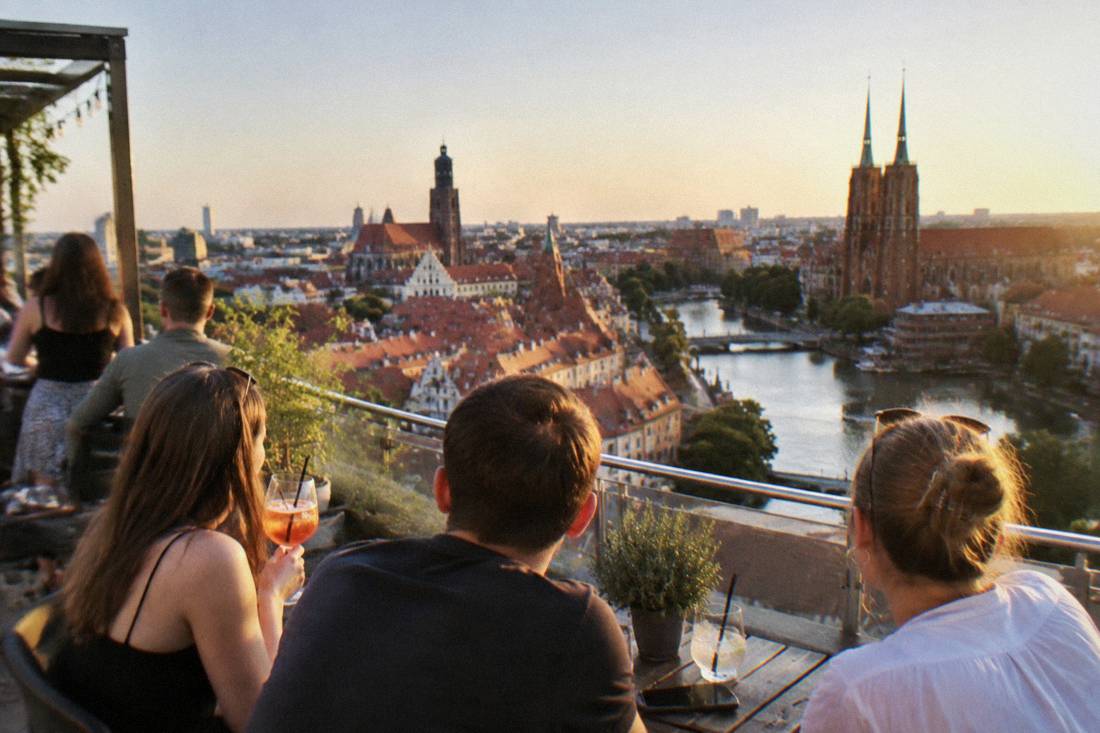 People with drinks at an open rooftop bar table in summer, overlooking Wroclaw rooftops in warm evening light.