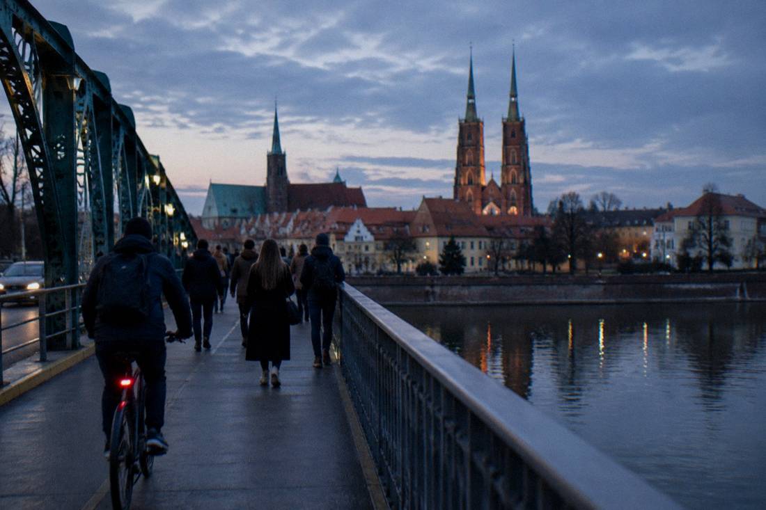 A cyclist and pedestrians crossing a bridge over the Odra river toward the Old Town, with Wroclaw church towers rising above the rooftops.