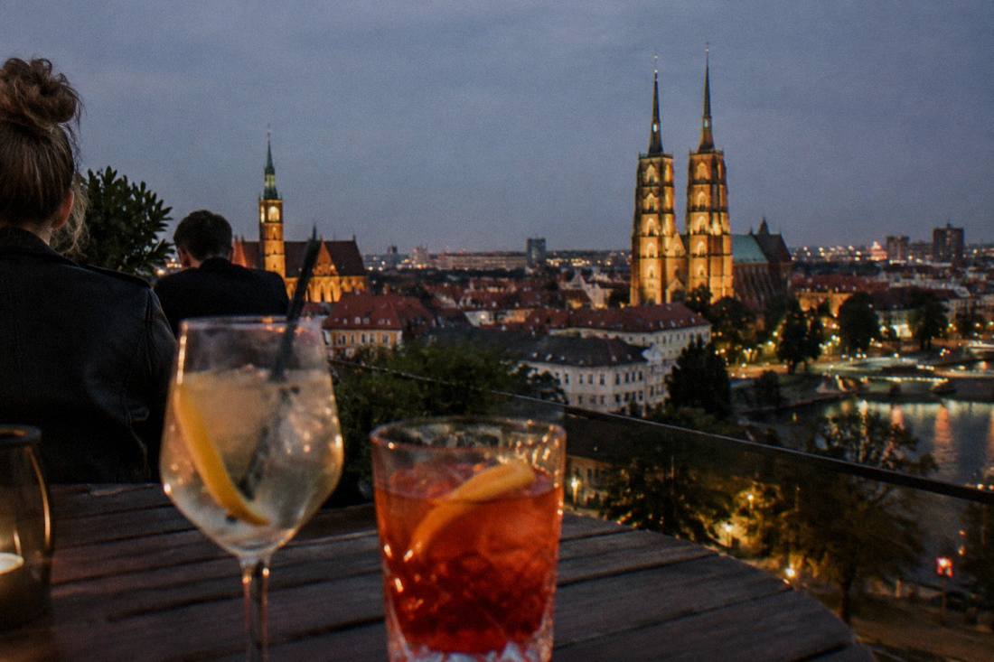 Close view of two drinks on a rooftop bar table with Wroclaw's illuminated church towers and dark rooftops in the background at night.