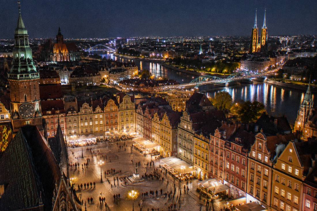 Wide night panorama of Wroclaw from above, showing the illuminated Market Square, the Odra river bridges and cathedral towers.