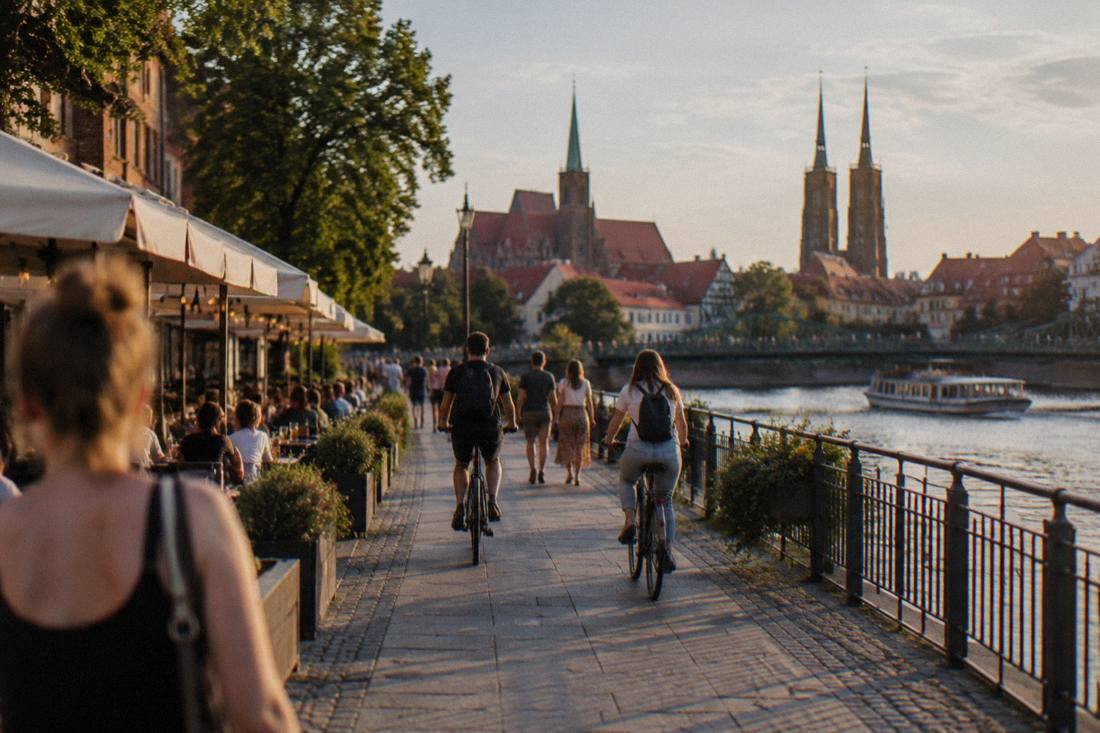 A riverside promenade with cyclists and pedestrians passing outdoor cafe terraces in late afternoon light