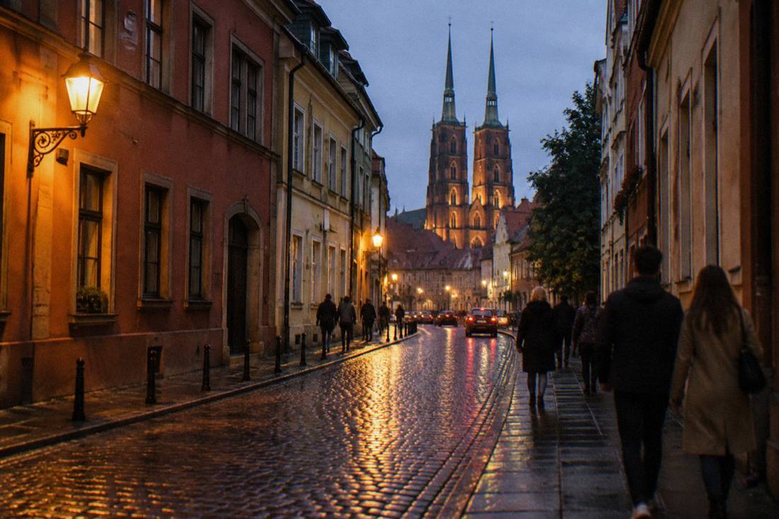 A narrow old town street at dusk with warm streetlights reflecting on wet cobblestones