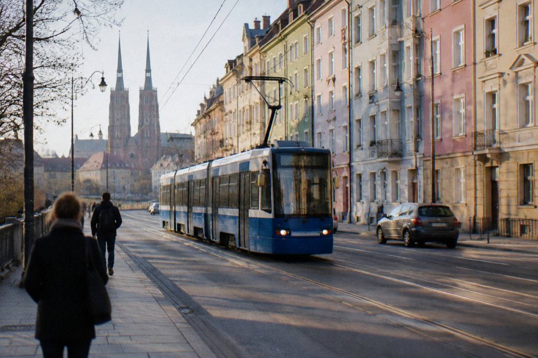 A tram moving along a city street lined with pastel-colored apartment buildings in daylight