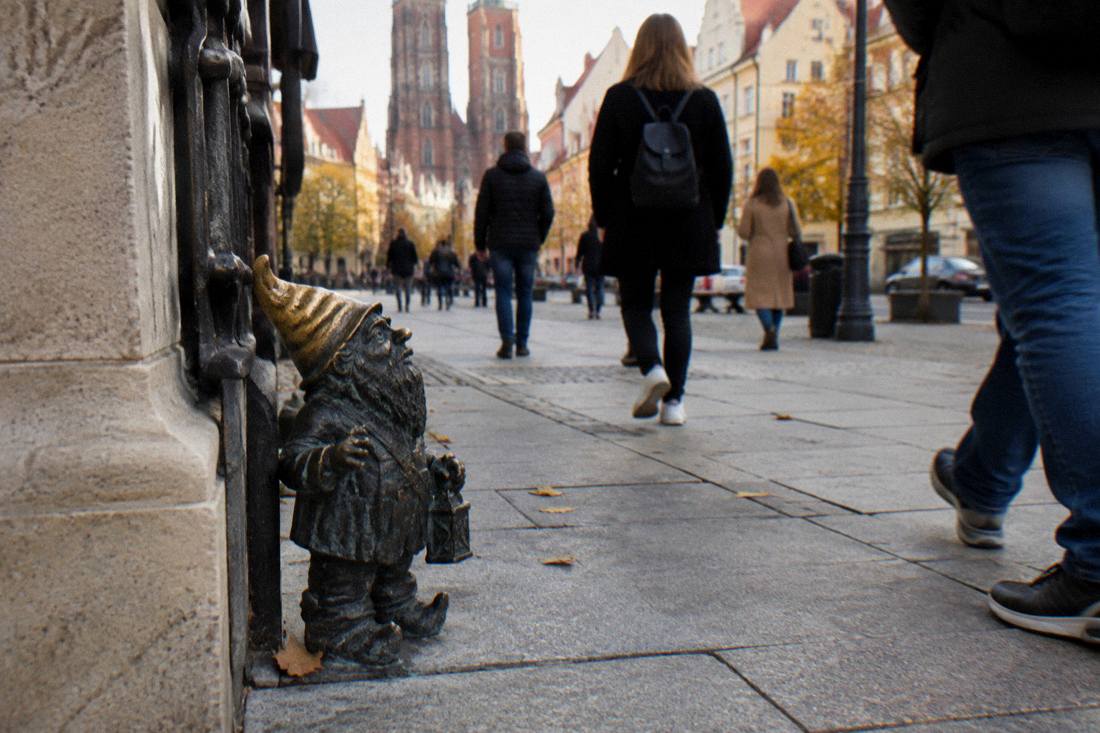 A small bronze dwarf statue standing on a pavement in central Wroclaw with people walking past