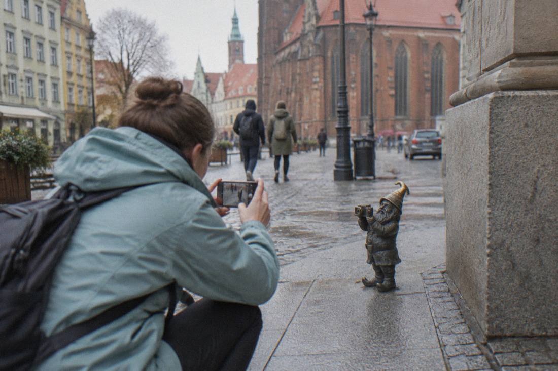 Visitor in a light rain jacket photographing a small bronze dwarf statue on a Wroclaw street