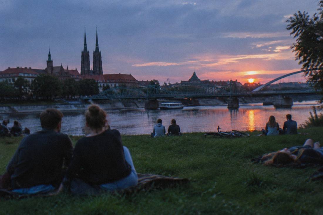 People relaxing at sunset on a grassy river island with Wroclaw's bridges in the background