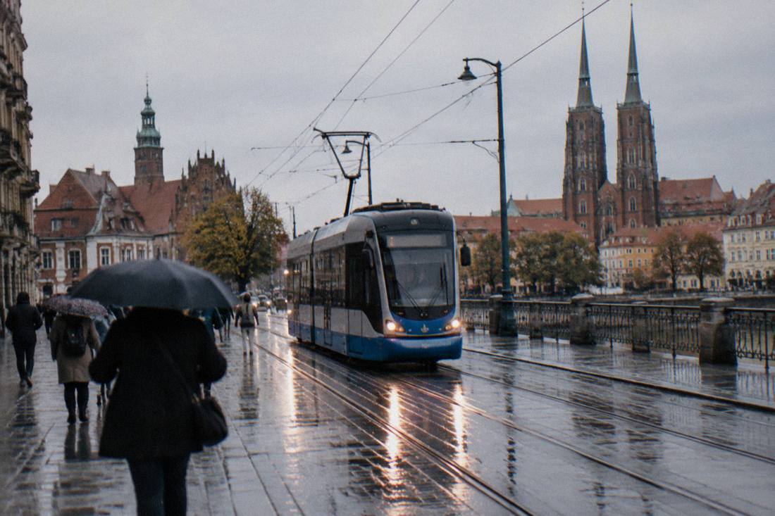 A modern tram on a wet city street with reflections on the rails and pedestrians with umbrellas