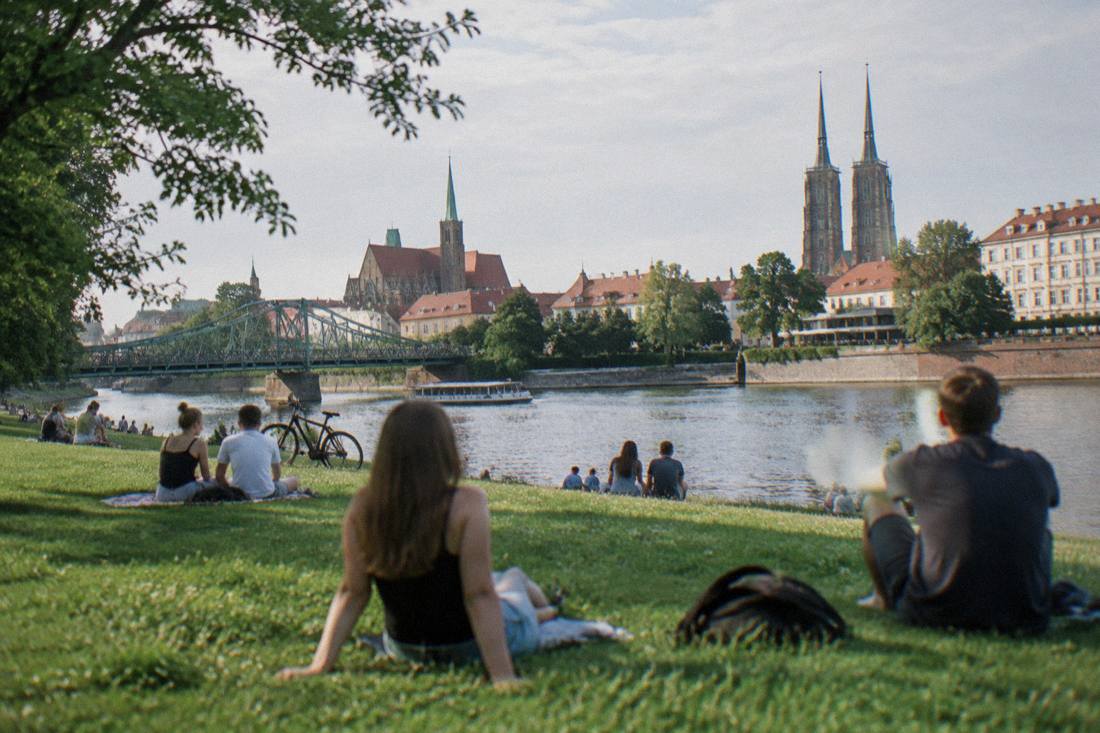 A green riverbank lawn on a Wroclaw island with people relaxing near the water in summer
