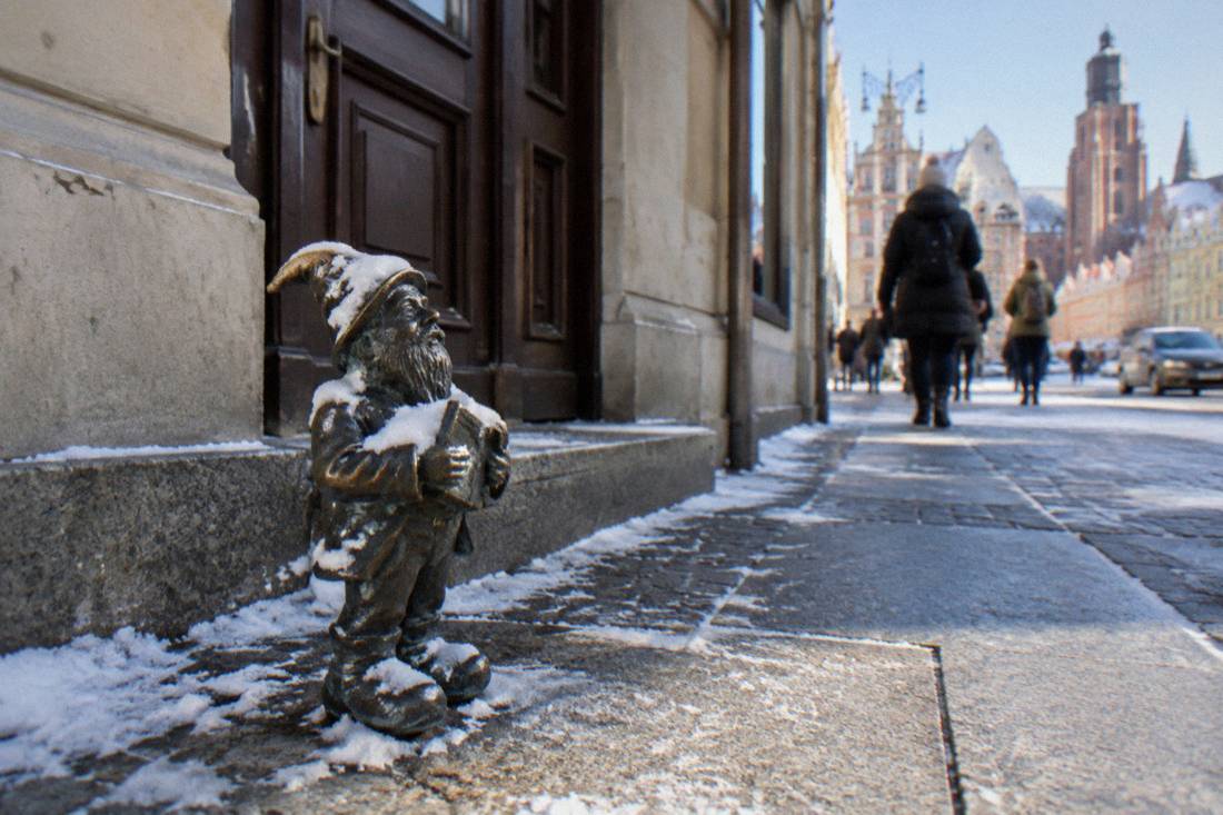 A small bronze dwarf statue on a Wroclaw sidewalk, dusted with snow, with a building doorway behind it.
